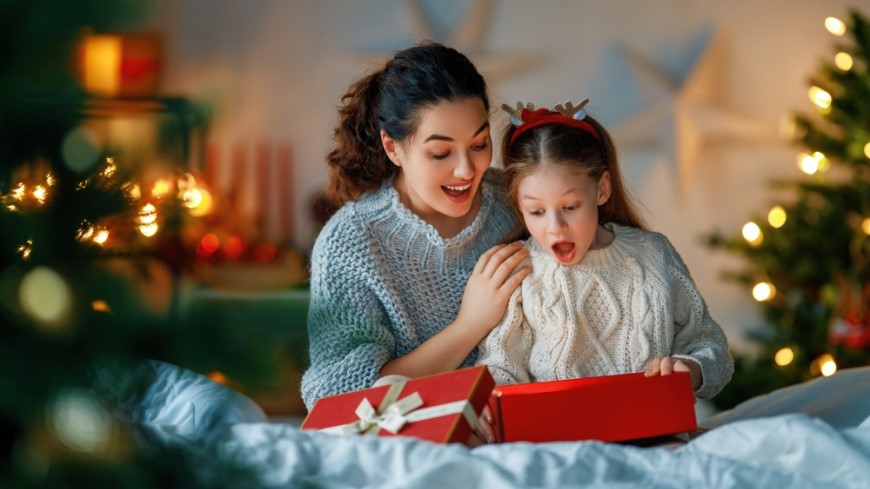 překvapená holčička si rozbaluje dárek u vánočního stromečku s maminkou