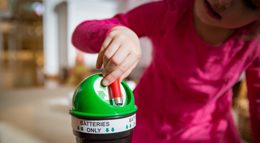 Holčička hází tužkovou baterii do malého koše se symbolem pro recyklaci,