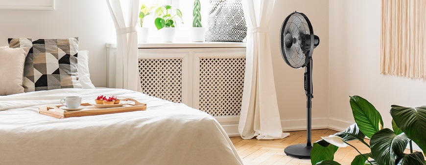 Bojujte s letními vedry s mobilními klimatizacemi ETA Elegantní černý stojanový větrák ETA Naos stojící v rohu světlé ložnice. V popředí je ustlaná manželská postel s podnosem s jídlem, v pozadí okno s bílými závěsy a pokojovými rostlinami