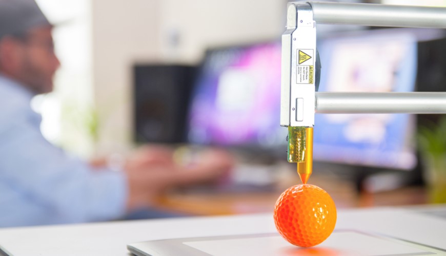 3D tiskárna vytváří oranžový golfový míček a v pozadí je muž pracující na pc