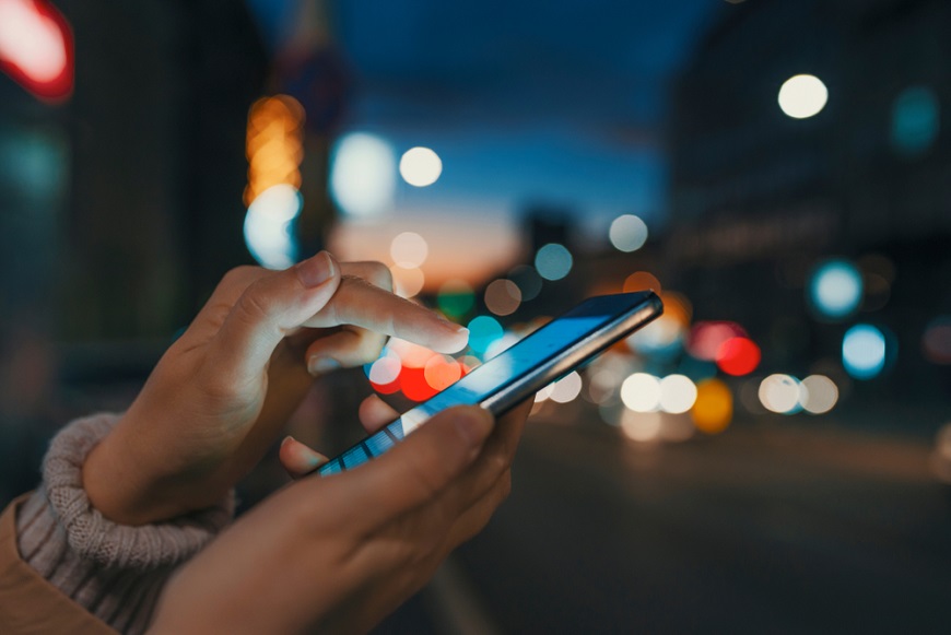 Jak restartovat mobil? detail rukou ovládajících chytrý telefon, v pozadí rušná ulice po setmění
