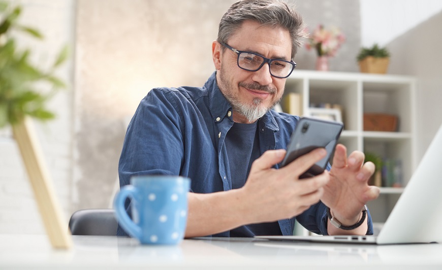 Jak restartovat mobil? muž sedí za stolem v pracovně a používá chytrý telefon, před sebou má otevřený notebook a šálek