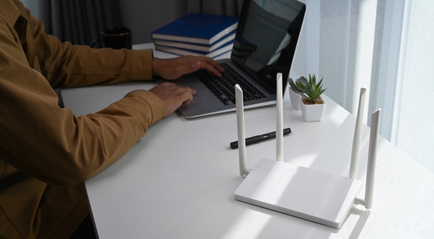 Jak propojit dva routery? Muž pracuje na MacBooku a vedle něho se nachází bílý Wi-Fi router