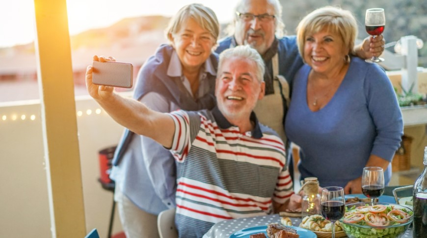 Starší muž se ženou a dalším starším párem pořizuje v restauraci u stolu selfie.