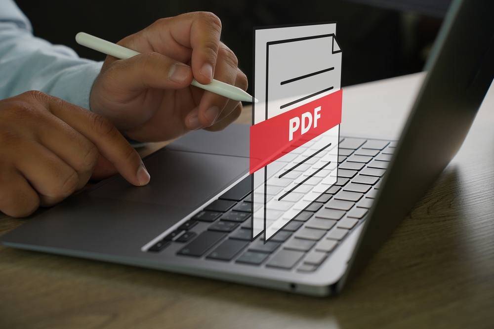 Muž drží jeden prst na touchpadu notebooku a v druhé ruce drží stylus. Nad klávesnicí notebooku je ikonu souboru pdf.