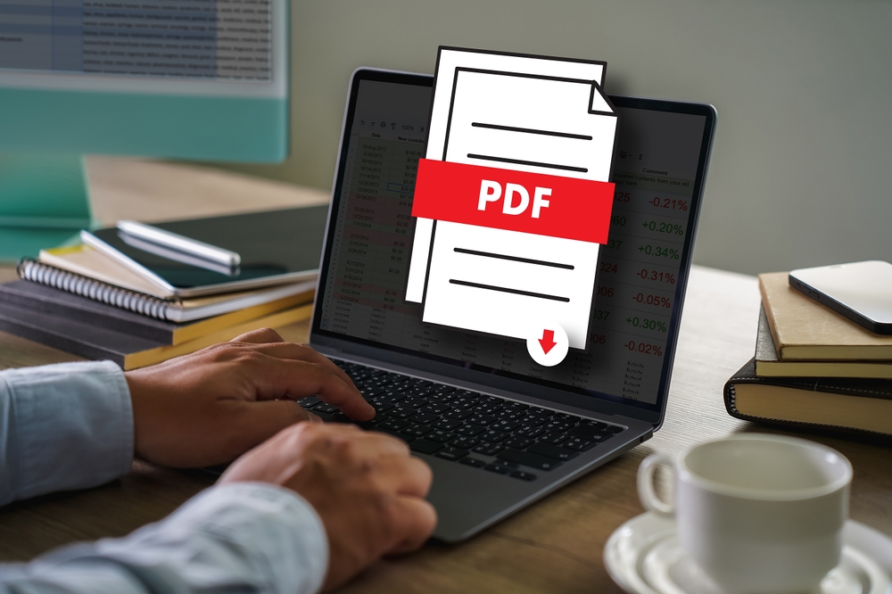 Notebook, přes jehož obrazovku je symbol staženého PDF souboru. Vlevo hromádka bloků a sešitů. Vpravo šálek od kávy a mobil na hromádce se sešity.