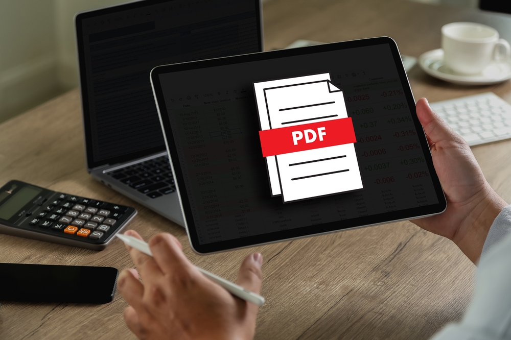 Muž drží tablet, na jehož obrazovce je symbol souboru PDF. V druhé ruce drží stylus. V pozadí je pracovní stůl s notebookem, kalkulačku, klávesnicí a šálkem kávy.