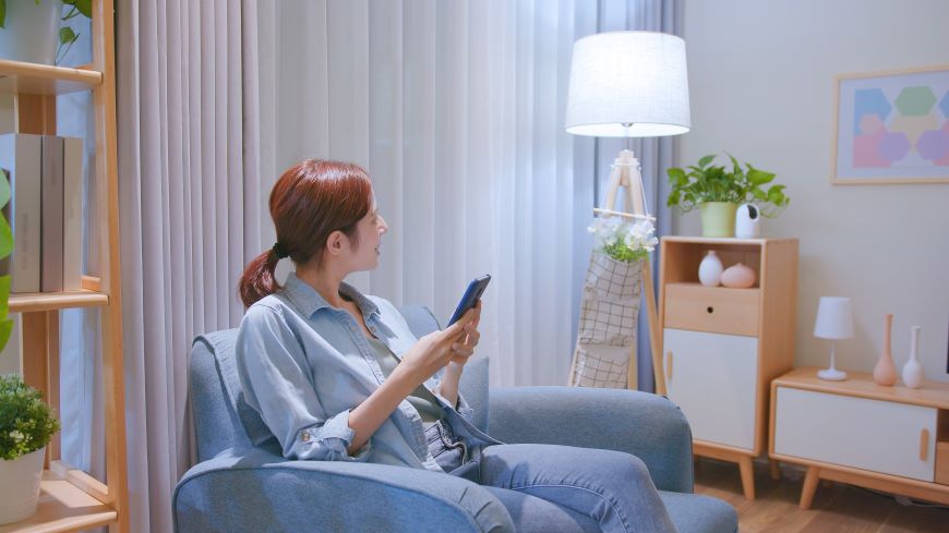 Jak ovládat televizi mobilem? Žena sedí v modrém křesle mezi nábytkem a hledí vpravo na lampu. Ta svití. Žena drží v ruce mobilní telefon – ovládá lampu pomocí mobilní aplikace.