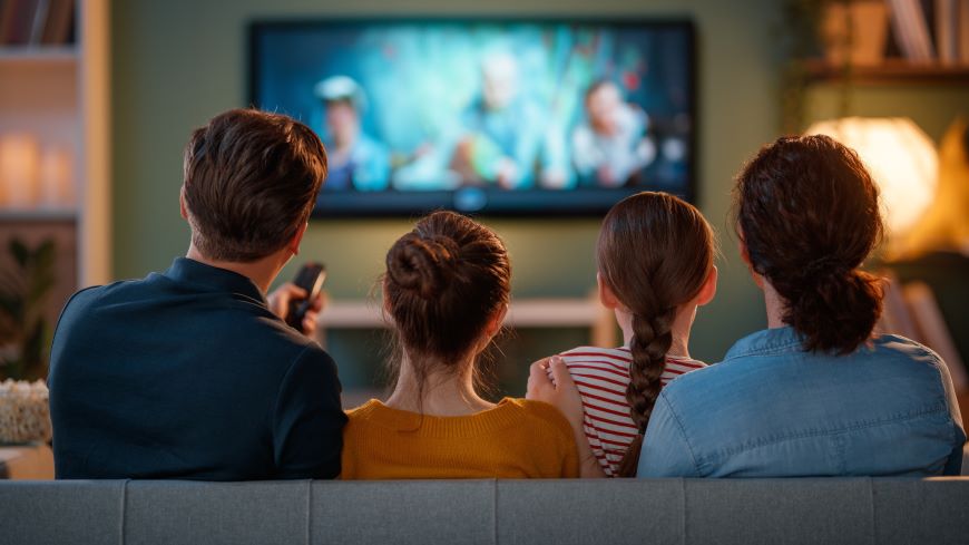 Jak ovládat televizi mobilem? Muž a žena se dvěma dětmi sedí před televizí – vidíme jejich záda. Muž ovládá televizi dálkovým ovladačem.