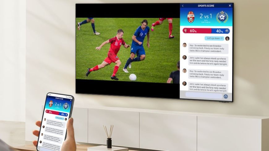Jak ovládat televizi mobilem? Mobilní telefon na jehož displeji jsou výsledky fotbalového zápasu. V pozadí je televize, na které je vlevo fotbalový zápas a vpravo totéž, co na displeji mobilního telefonu.