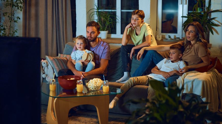 Jak ovládat televizi mobilem? Na pohovce sedí žena s mužem se syny a dcerou Na stole mají pití a popcorn a hledí na zapnutou televizi.