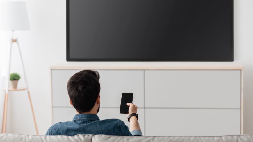 Jak ovládat televizi mobilem? Muž sedí na pohovce před vypnutou televizí a míří na ni mobilním telefonem.