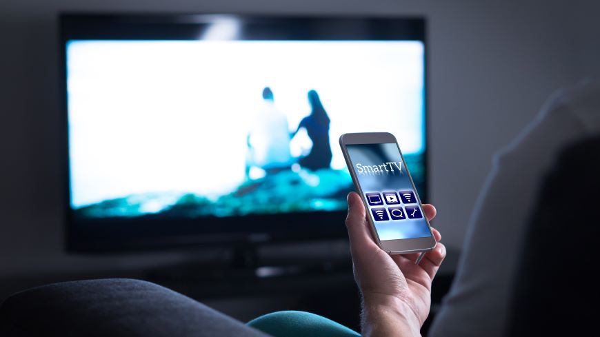 Jak ovládat televizi mobilem? Mobilní telefon s aplikací Smart TV na displeji. V pozadí zapnutá televize.