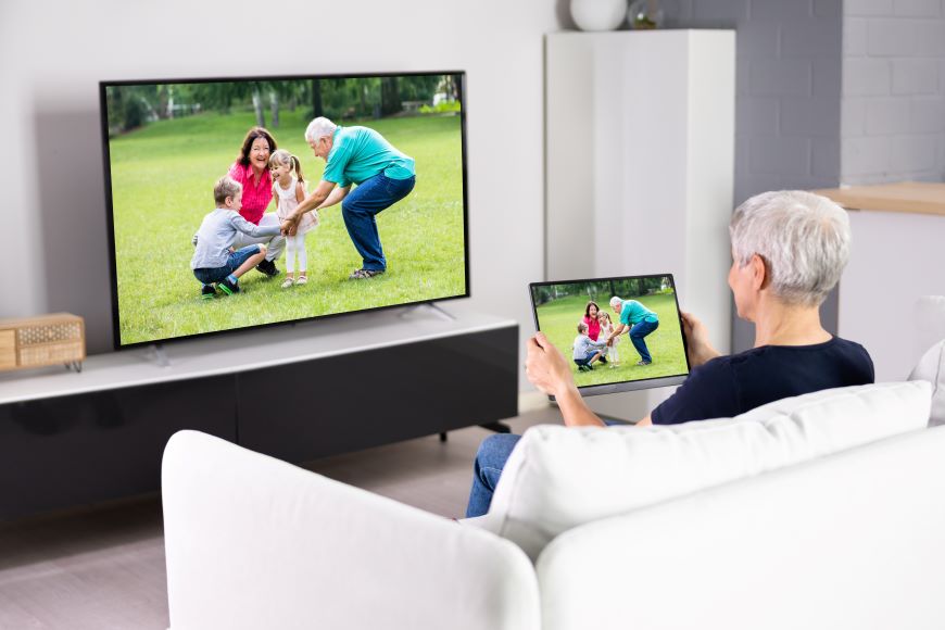Starší žena sedí na pohovce před TV a dívá se na tablet, který drží v rukou. Na tabletu je stejná fotografie rodiny s dětmi jako na TV.