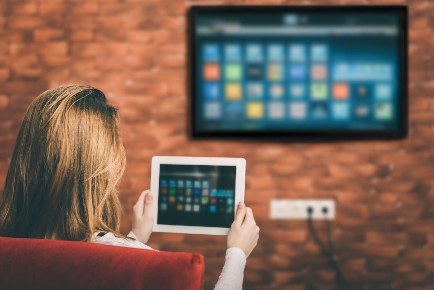 Žena sedí na pohovce před televizí a drží před sebou tablet. Na TV se zrcadlí totéž, co je na displeji tabletu.
