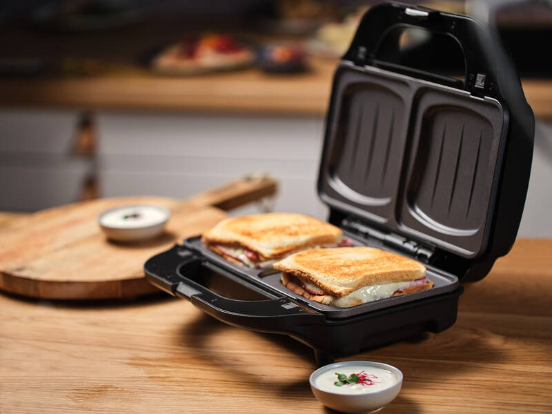 TOP 10 nejlepších dárků značky ETA Otevřený černý sendvičovač ETA Sorento Plus s hotovými zapečenými toasty se šunkou a sýrem, umístěný na dřevěné kuchyňské desce