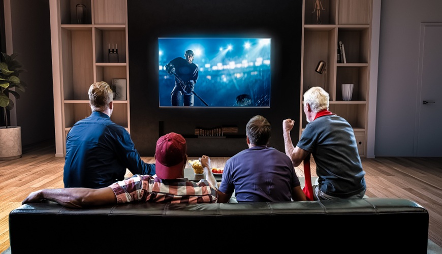 Jak si užít sport na maximum aneb Průvodce elektronikou pro opravdové fanoušky Kvarteto přátel, sedící na gauči v obývacím pokoji, sleduje hokejový přenos na televizní obrazovce.