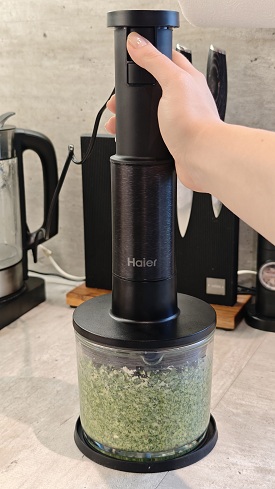 ruka drží tyčový mixér Haier I-Master Series 5, který je připojený k sekáčku, uvnitř se tvoří pesto