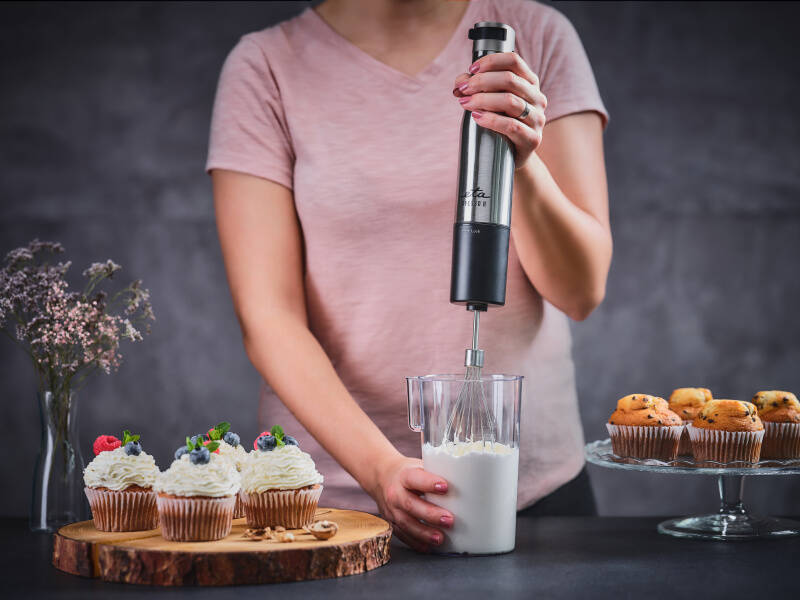 Žena v růžovém tričku používá nerezový tyčový mixér ETA Spesso II se šlehací metlou k přípravě krému v odměrné nádobě, vedle jsou nazdobené borůvkové cupcakes.