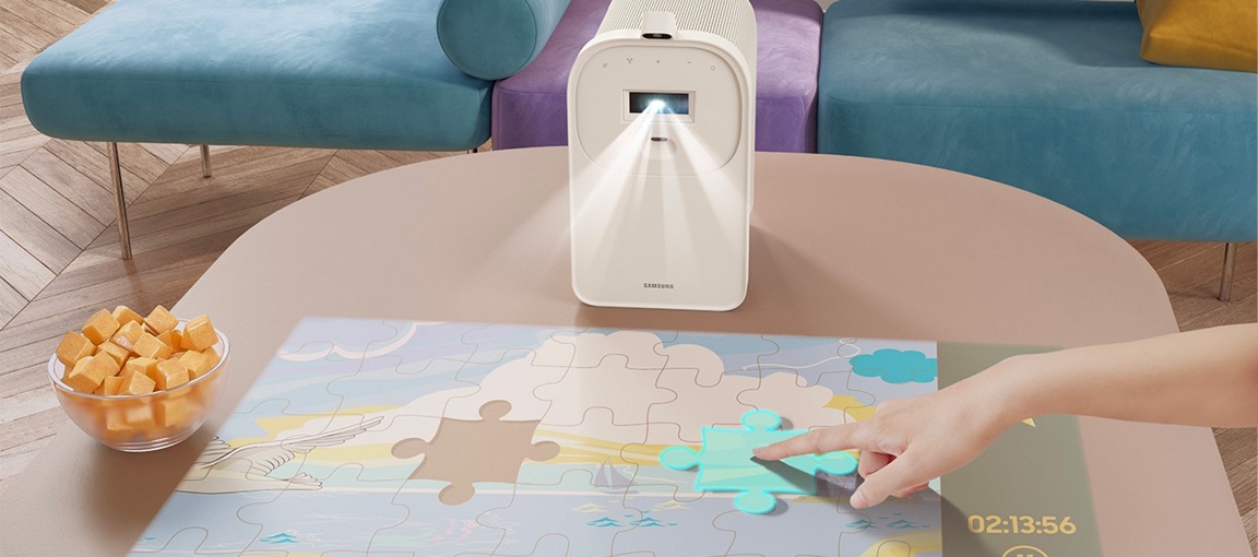 Lifestylová kolekce Samsung: Designové televizory a soundbary, které zkrášlí váš domov Projektor Samsung The Premiere 5 ve stojánku promítá aplikaci s puzzle na stůl, lidská ruka posouvá pomocí dotykového ovládání dílek puzzle