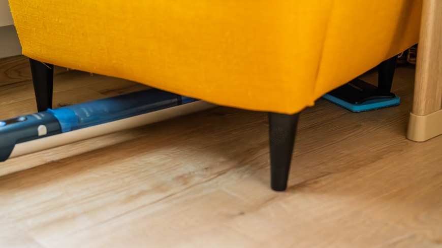 Recenze: Elektrický mop Philips OneUp – vytírejte jedním tahem a bez námahy Mop Philips OneUp zasunutý pod žlutým křeslem – položený na podlaze.