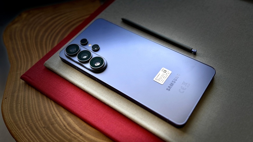 Smartphone Samsung Galaxy S26 Ultra ve fialovém provedení spolu s perem S pen a zátiším v podobě vysokoškolských diplomů, navrstvených na sobě