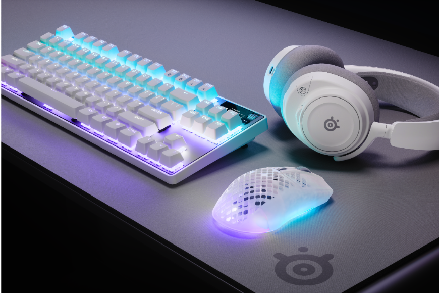 Nová myš SteelSeries Aerox 3 Wireless Gen 2: Vylepšená generace je tady Bílá myš SteelSeries Aerox 3 Wireless Gen 2 a modrofialovým podsvícením leží na stole vedle headsetu a klávesnice SteelSeries