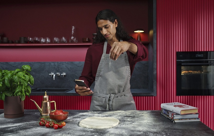 Nová trouba AEG PizzaExpert: Pizza jako z Neapole jen za 2,5 minuty Muž připravuje pizzu a dívá se do mobilního telefonu. V pozadí je červená kuchyň s troubou AEG.
