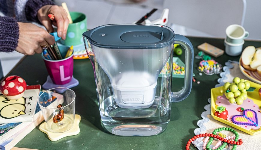Filtrační konvice Brita Style Essential XL v tmavě modré barvě umístěna na stole s pastelkami, hrníčky apod.