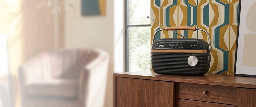 Retro rádio JVC RA-E215G v černo-dřevěném provedení s kovovým madlem, umístěné na dřevěné komodě před výraznou vzorovanou tapetou