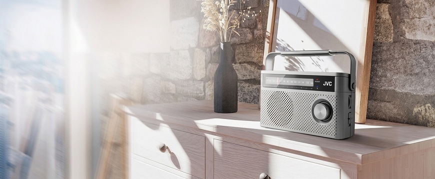 Stříbrné přenosné rádio JVC RA-E315 na světlé dřevěné komodě zalité sluncem, umístěné před kamennou stěnou s dekoracemi