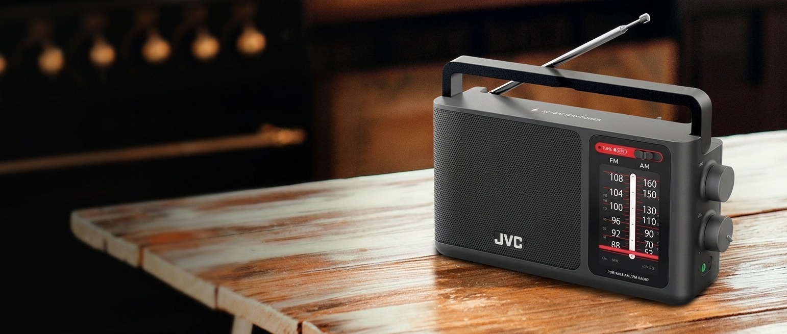 Detailní záběr na černé analogové rádio JVC RA-E435B  s AM/FM laděním. Rádio má integrované madlo a stojí na masivním dřevěném povrchu, v pozadí je rozostřená kuchyňská linka