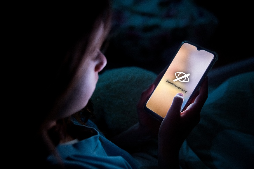 Jak nastavit rodičovskou kontrolu na chytrých zařízeních? Dívka leží v noci posteli a do obličeje jí svítí displej mobilu, na kterém je symbol pro sensitive content.