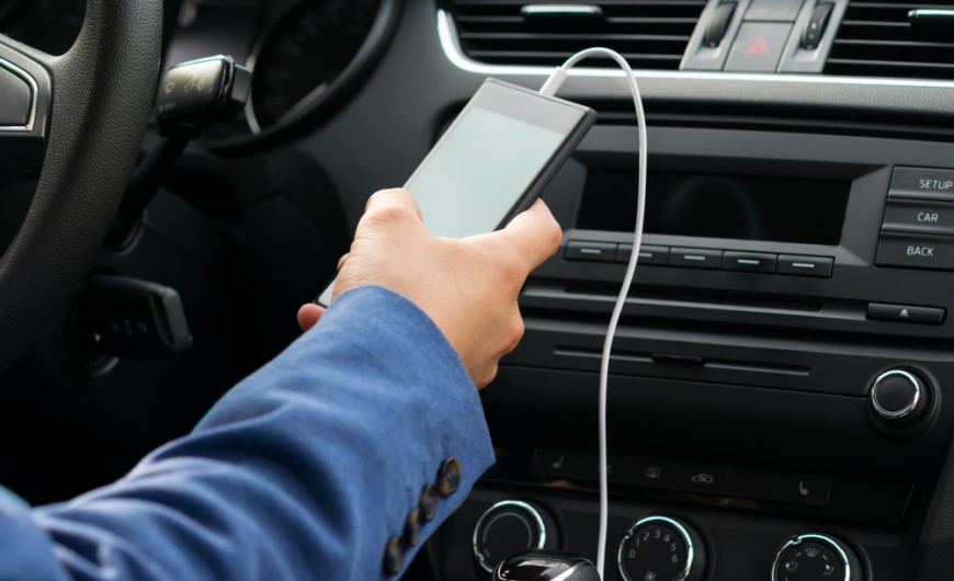 Jak spárovat telefon s autem: Průvodce, se kterým to zvládnete za 2 minuty Muž sedí v autě na místě řidiče a má připojen chytrý telefon přes kabel k autu
