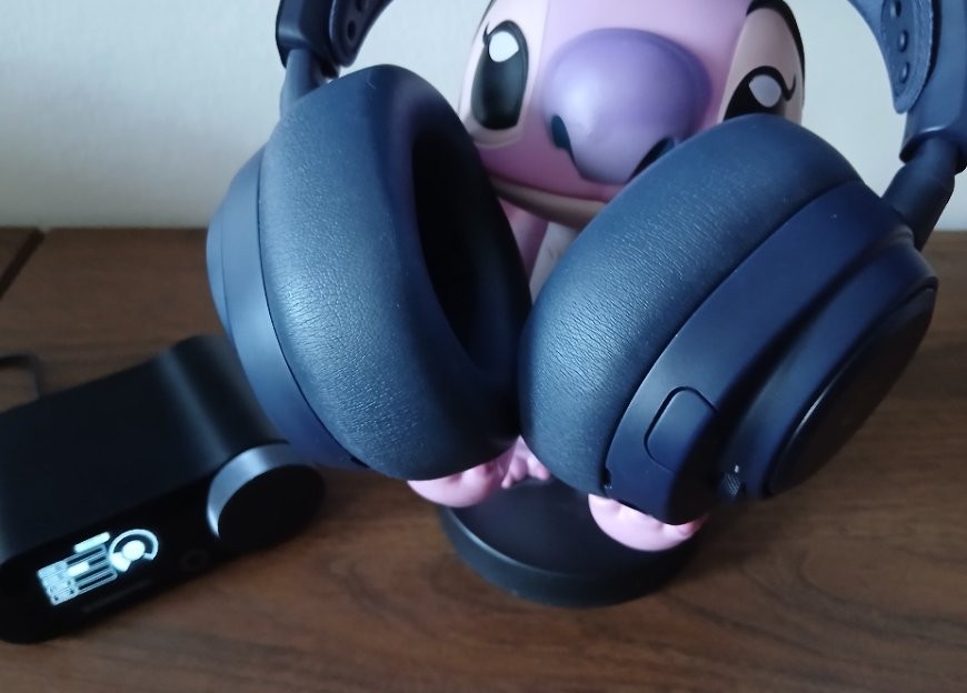 Headset Arctis Nova Pro Omni je položený na stojánku ve tvaru postavičky, vedle leží zapnutý GameHub