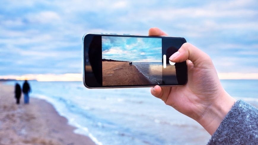 Jak pořídit dobré fotky mobilem? Smartphone fotografující pár v pozadí, jdoucí po pláži vedle možského příboje.