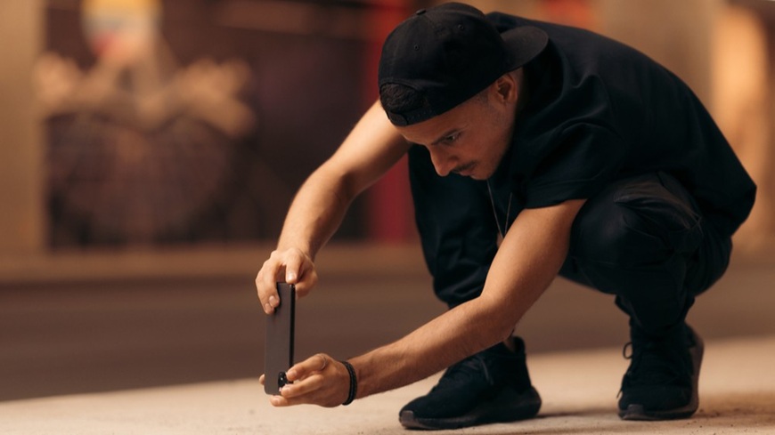 Jak pořídit dobré fotky mobilem? Fotografování z žabí perspektivy – černě oděný muž v podřepu ostřící smartphonem na blízký objekt.