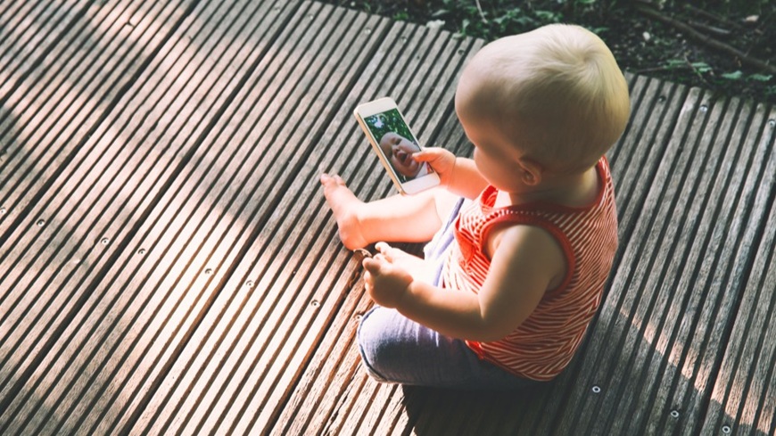 Jak pořídit dobré fotky mobilem? Batole sedící na verandě a držící v ruce mobilní telefon se spuštěnou fotoaplikací