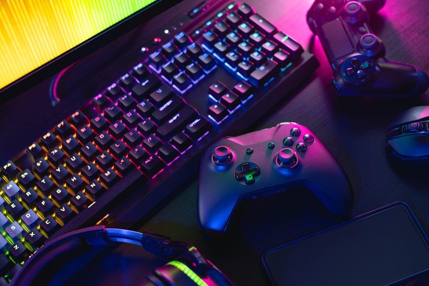 Prázdniny 2026 hrou: Odměňte gamera za vysvědčení! Na stole leží před monitorem klávesnice, 2 gamepady a myš