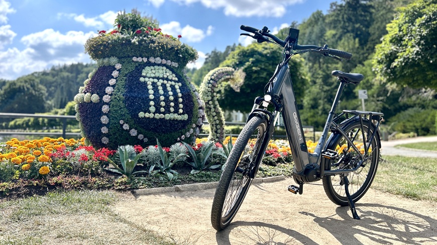Recenze: Elektrokolo GoGEN Aspen MX 720 se postará o to, aby vaše cyklochoutky nic nebrzdilo Elektrokolo GoGEN Aspen MX 720 v šedo-černém provedení opřené o stojan před prosluněným kvetoucím záhonem