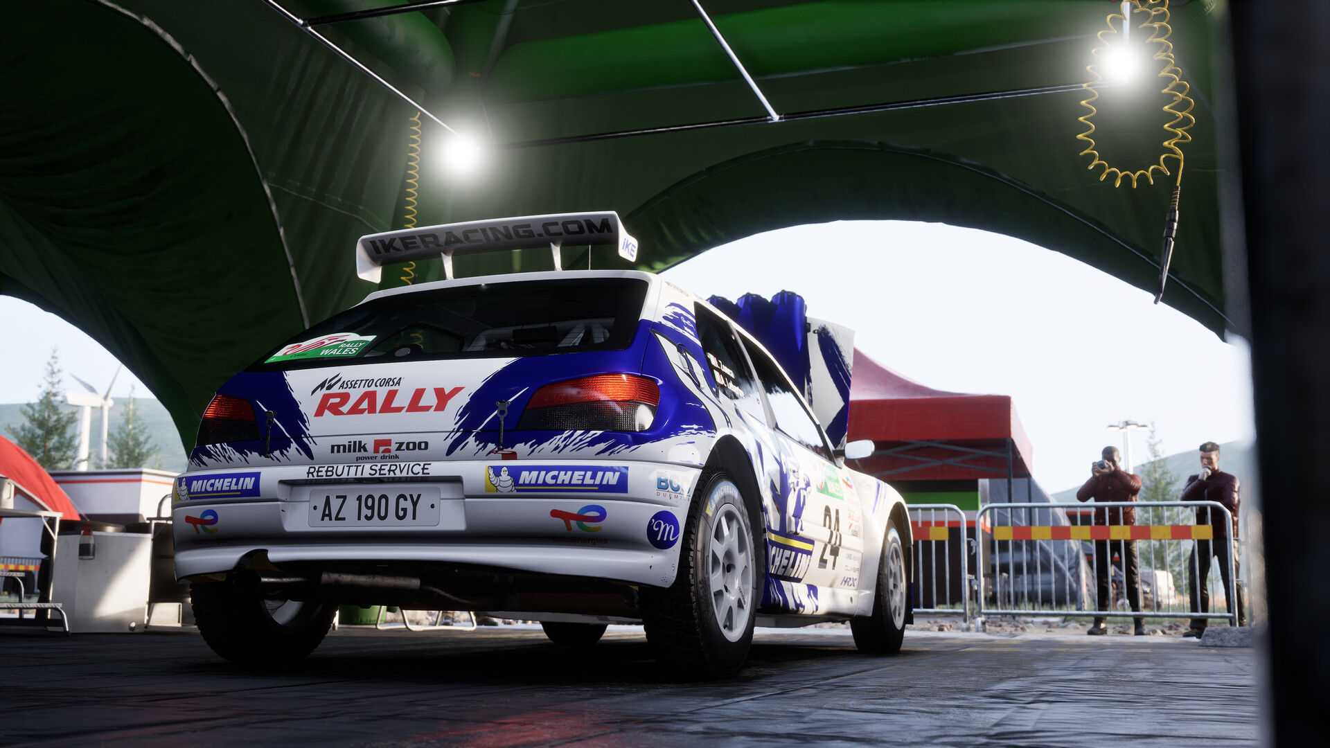 Assetto Corsa Rally: Šampion v simracingovém realismu Pohled na závodní auto zezadu, před autem stojí dva lidé