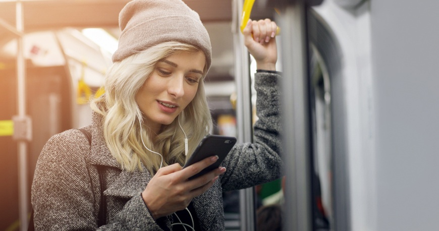 Žena v zimním oblečení poslouchá hudbu v tramvaji přes sluchátka a dívá do chytrého telefonu