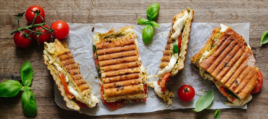 Grilované panini s mřížkou plněné mozzarellou, rajčaty a bazalkovým pestem, nakrájené na pečicím papíře na dřevěném stole. Příprava v sendvičovači ETA Sorento.