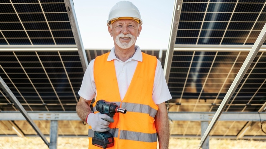 Jak na fotovoltaiku v roce 2026: Získejte solární elektrárnu bez úroků a počátečních úspor! Muž důchodového věku s utahovákem v ruce, chystající se na montáž/revizi solárních panelů v pozadí.