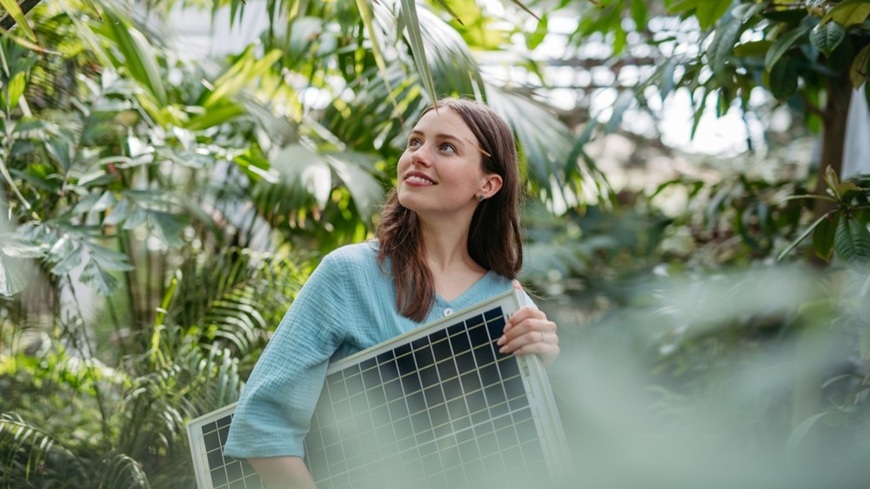 Jak na fotovoltaiku v roce 2026: Získejte solární elektrárnu bez úroků a počátečních úspor! Žena se solárním panelem v podpaží, pozorující rozkvetlou zeleň v okolí.