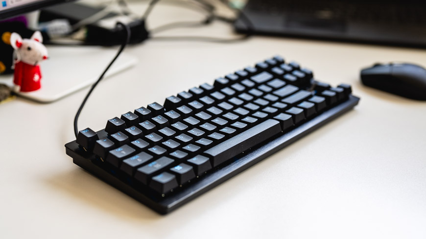 Herní klávesnice Razer Huntsman Tenkeyless V3 8KHz leží na stole, na pozadí je počítačová myš, stojan monitoru a figurka myšky