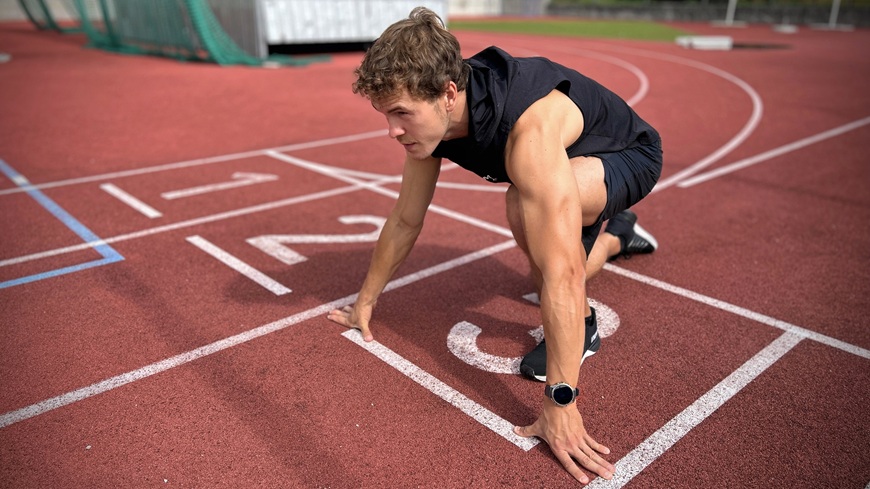 Muž s chytrými hodinkami Garmin fēnix® 9 Pro na zápěstí se v bloku na atletickém ovále připravuje na sprint.