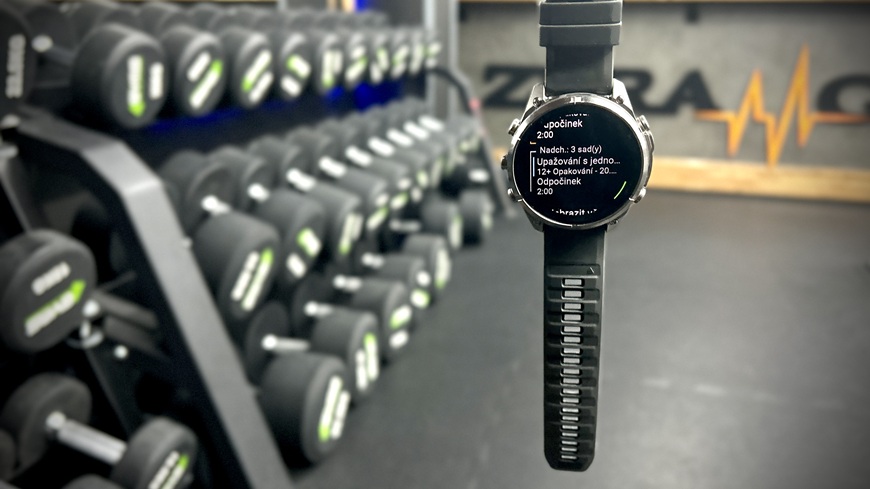 Chytré hodinky Garmin fēnix® 9 Pro visící v popředí stojanu s činkami v posilovně
