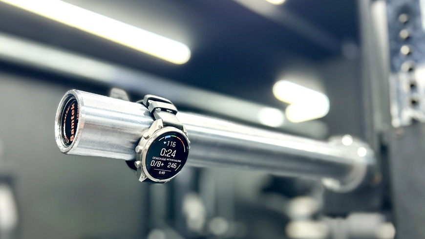Chytré hodinky Garmin fēnix® 9 Pro zavěšené na olympijské vzpěračské ose v posilovně.
