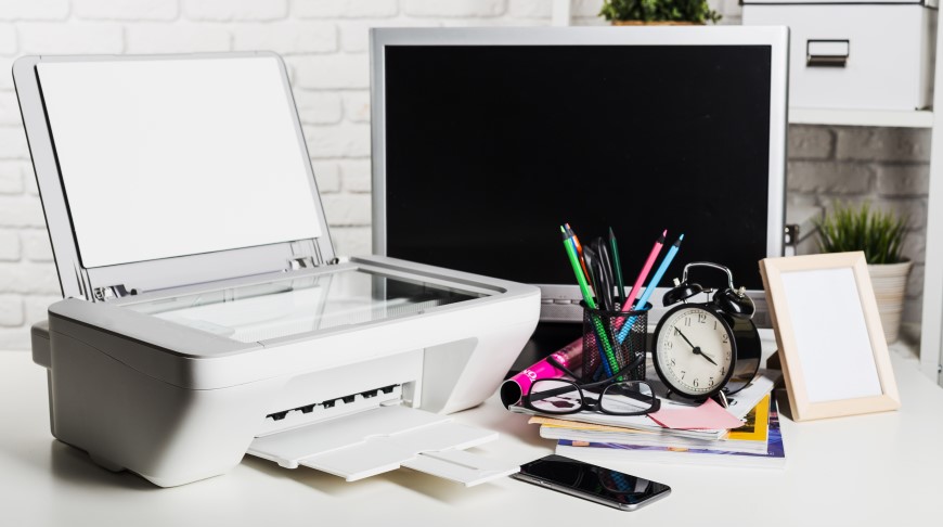Jak vybrat tiskárnu? Tiskárna na psacím stole. Vedle je monitor a psací potřeby s budíkem.